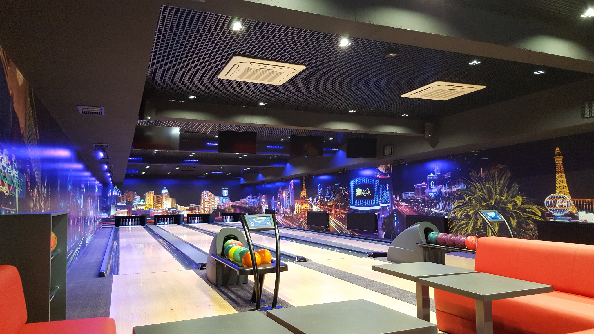 Nowoczesne wnętrze kręgielni z neonowym oświetleniem, automatami do podawania kul i fototapetą z panoramą miasta. Widoczne stoliki i sofy dla gości.