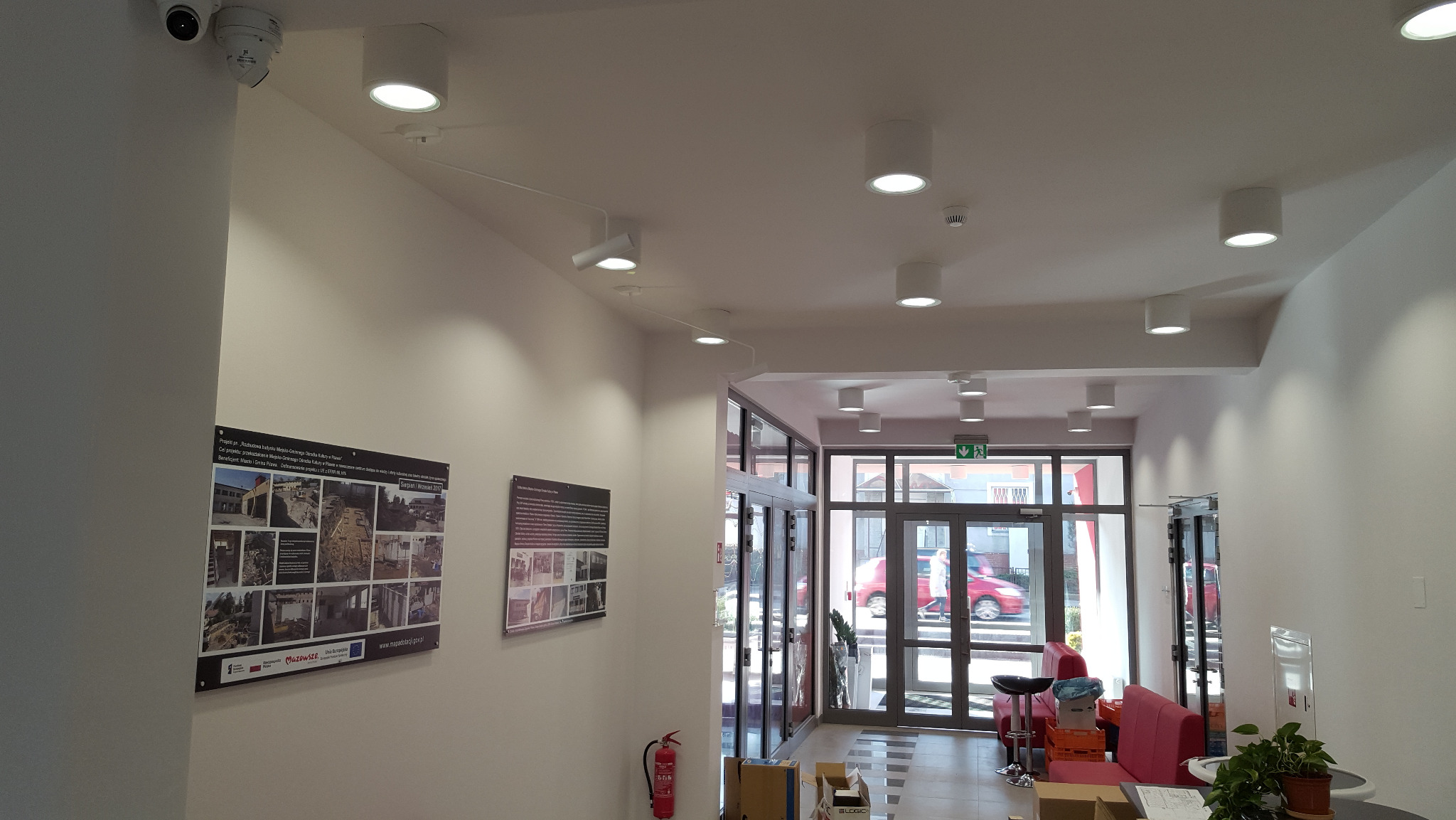 Białe, okrągłe lampy sufitowe w jasnym holu budynku, z widocznymi plakatami na ścianie i kamerą monitoringu. Nowoczesne oświetlenie w przestrzeni publicznej.