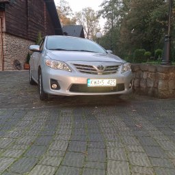 TransDave Dawid Tatka - Srebrna Toyota Corolla na tle kamiennego budynku i zieleni. Widoczna tablica rejestracyjna. Ujęcie z dołu, podkreślające bryłę auta.