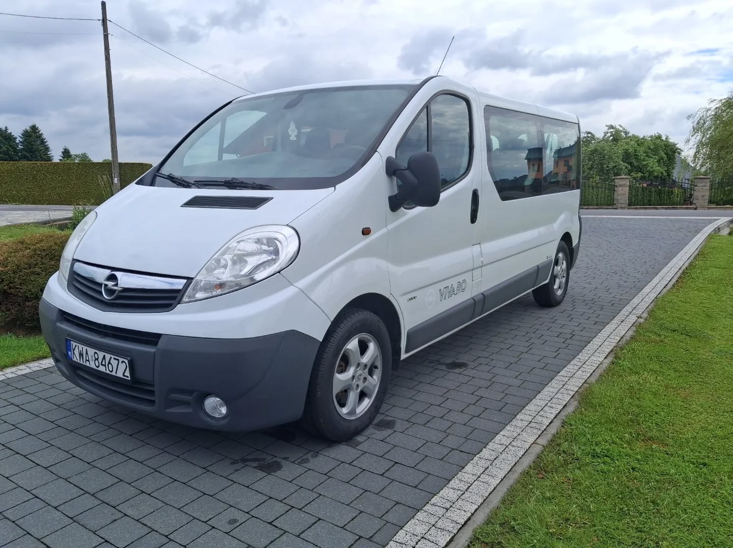 Biały Opel Vivaro z anteną na dachu, zaparkowany na szarym bruku. Widoczne tablice rejestracyjne i zielone otoczenie. Auto osobowe z szybami.
