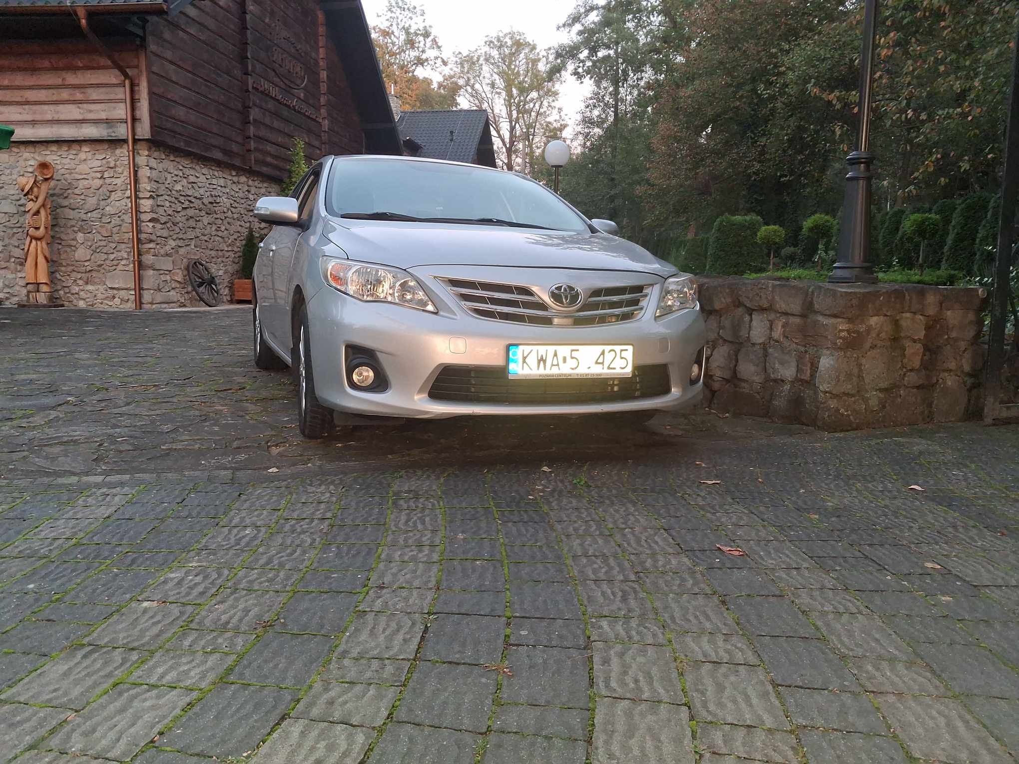 Srebrna Toyota Corolla na tle kamiennego budynku i zieleni. Widoczna tablica rejestracyjna. Ujęcie z dołu, podkreślające bryłę auta.