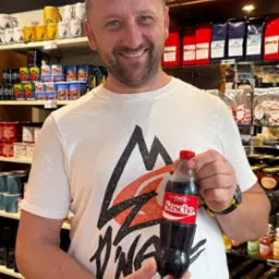 Uśmiechnięty mężczyzna trzyma w dłoni spersonalizowaną butelkę Coca-Coli z imieniem 'Sascha' na etykiecie, na tle półek sklepowych z pamiątkami i słodyczami.