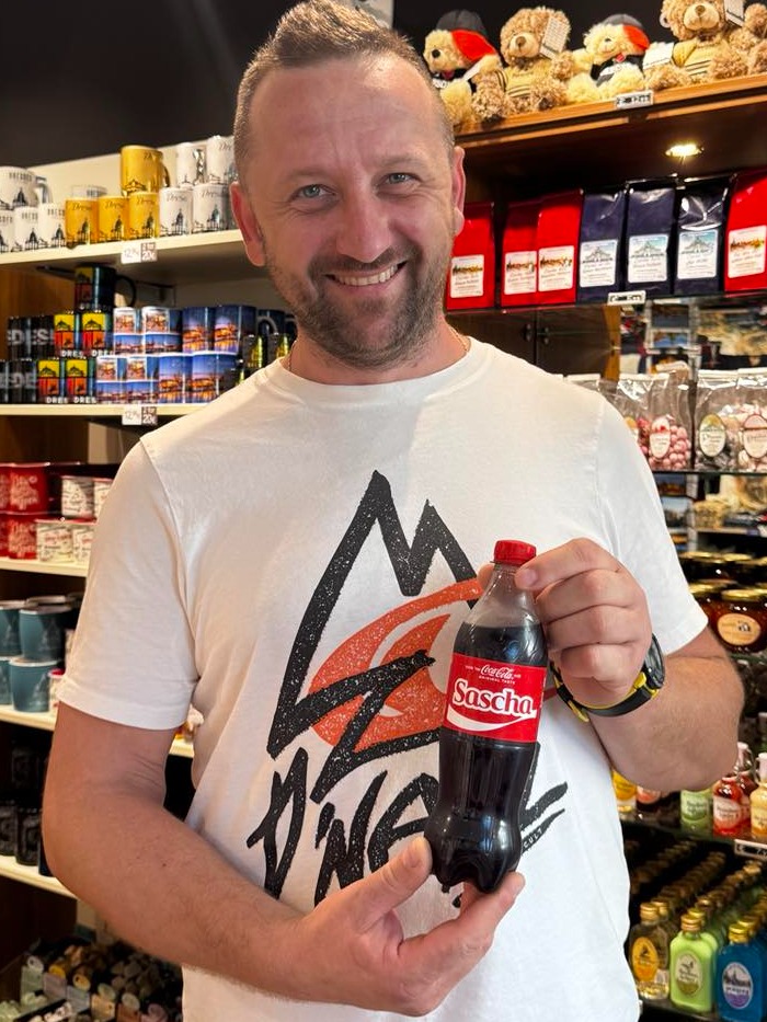 Uśmiechnięty mężczyzna trzyma w dłoni spersonalizowaną butelkę Coca-Coli z imieniem 'Sascha' na etykiecie, na tle półek sklepowych z pamiątkami i słodyczami.