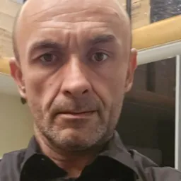 Portret mężczyzny z bliska, widoczne zmarszczki na czole, krótko ostrzyżone włosy, ciemna koszula, neutralny wyraz twarzy, w tle fragment okna i kartonów.