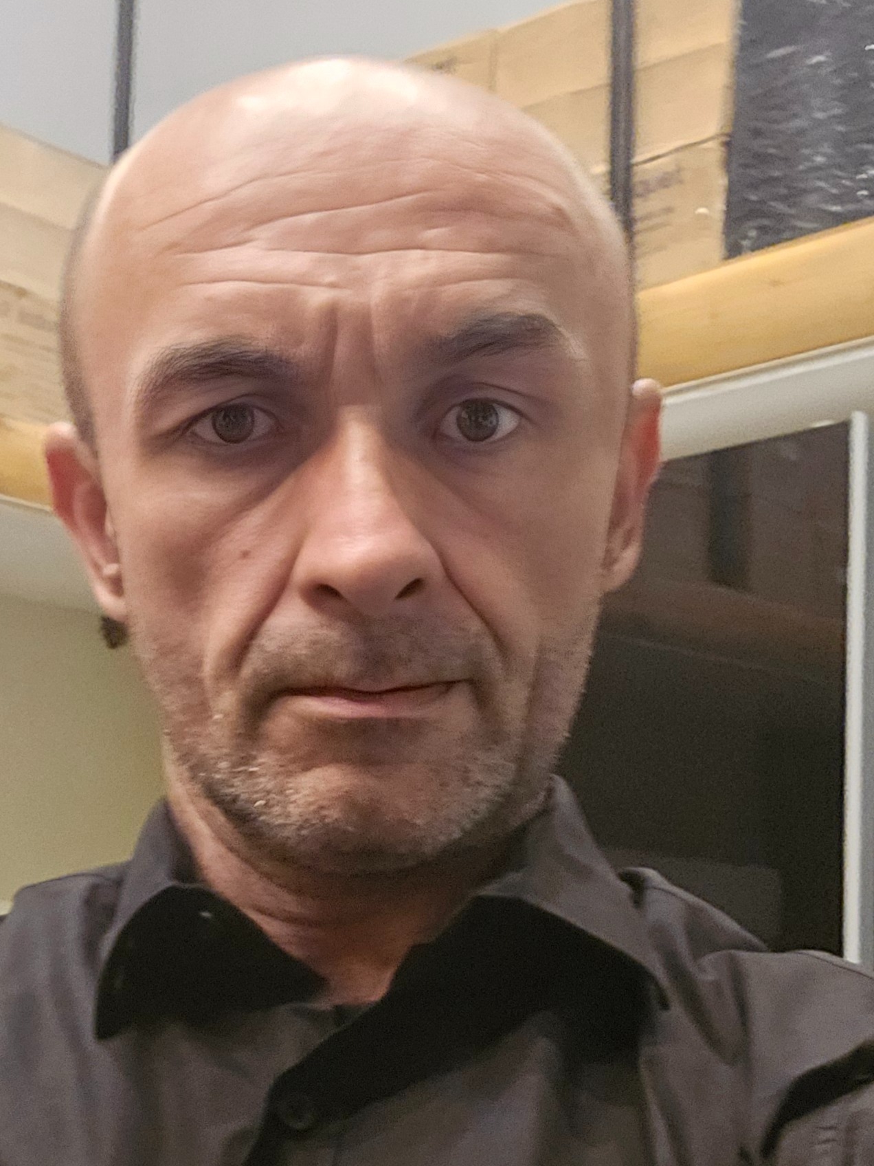 Portret mężczyzny z bliska, widoczne zmarszczki na czole, krótko ostrzyżone włosy, ciemna koszula, neutralny wyraz twarzy, w tle fragment okna i kartonów.