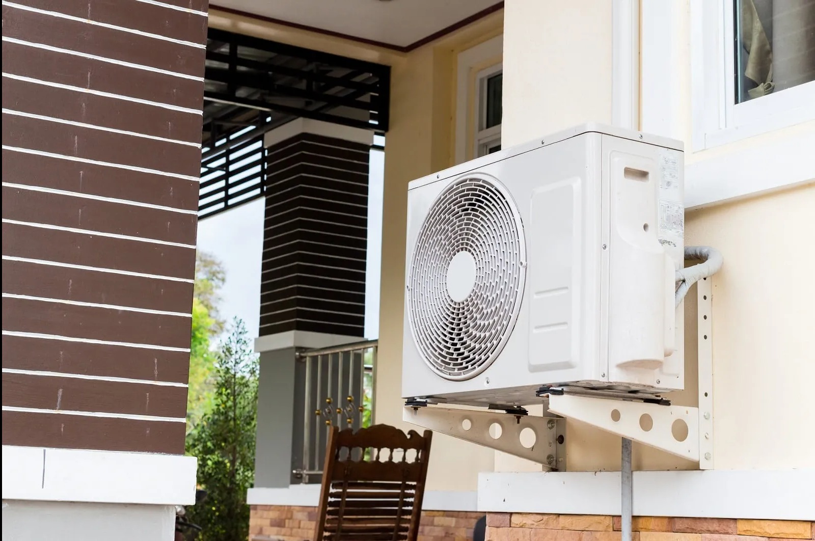 Biała jednostka zewnętrzna klimatyzacji zamontowana na elewacji domu z beżową ścianą i brązowymi elementami dekoracyjnymi. Widoczny fragment okna i krzesła.