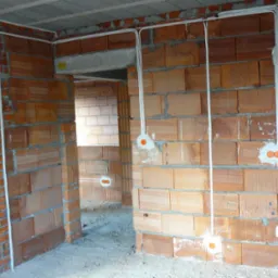 Surowe mury z instalacją elektryczną: białe przewody prowadzone po cegłach, pomarańczowe puszki. Widok wnętrza w trakcie prac, z betonowym nadprożem.