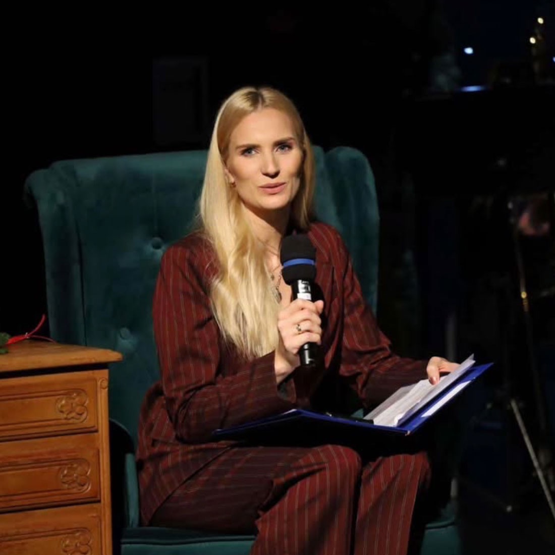 Blond prezenterka w bordowym garniturze w paski, siedzi w fotelu z mikrofonem i dokumentami, prowadząc wydarzenie na scenie w ciemnym otoczeniu.