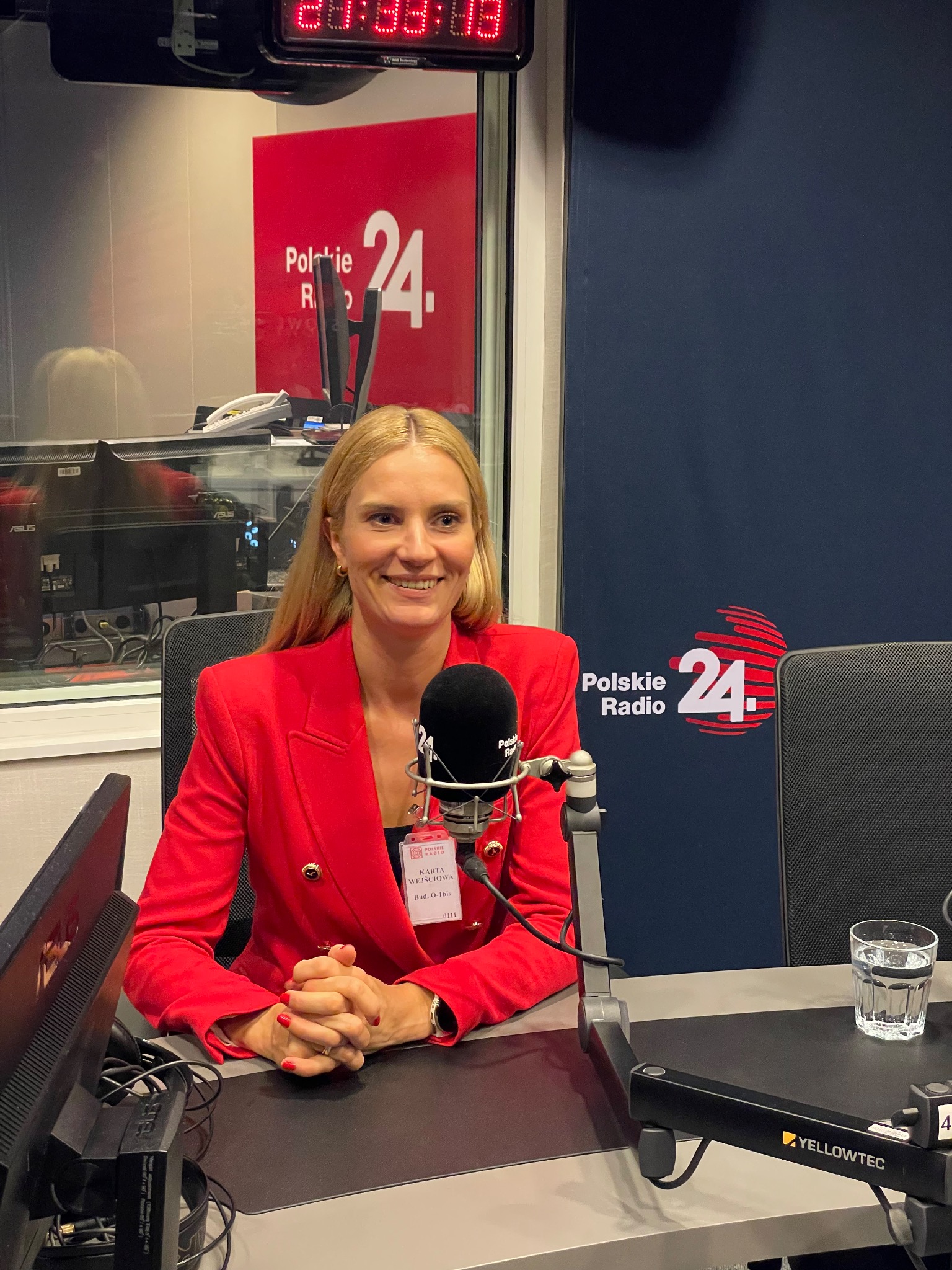 Kobieta w czerwonej marynarce siedzi w studiu Polskiego Radia 24, uśmiechając się do kamery. Na biurku mikrofon i szklanka wody.