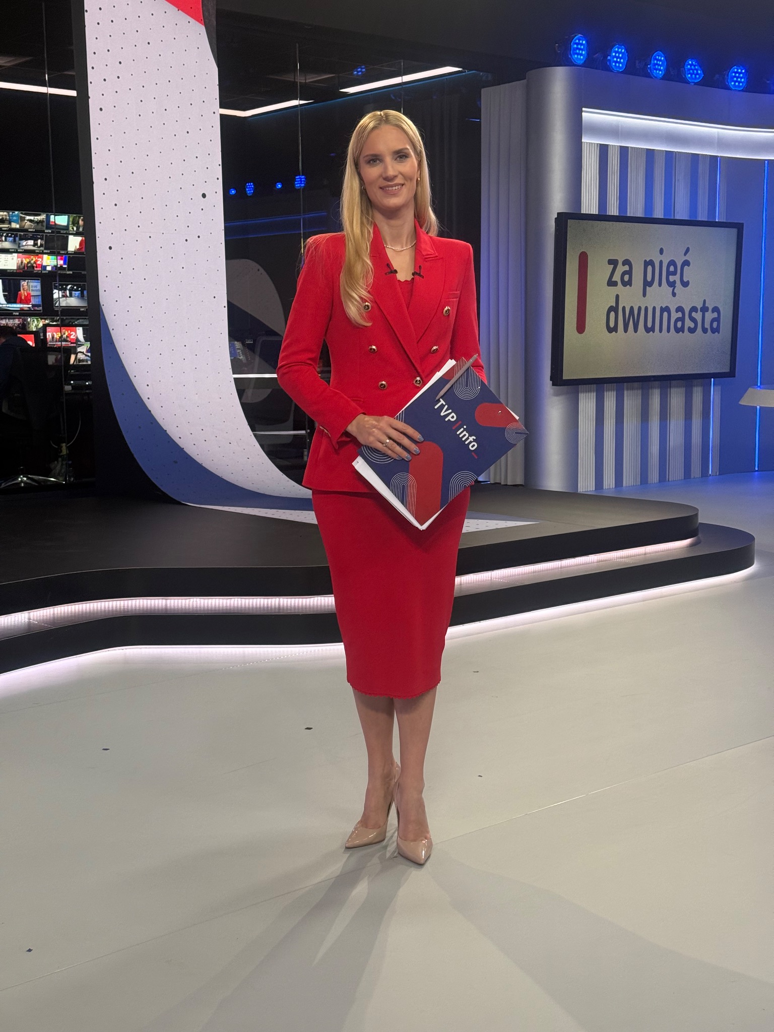 Prezenterka w czerwonym kostiumie na planie studia telewizyjnego, trzymająca w ręku materiały TVP Info. W tle scenografia z oświetleniem LED i ekran z napisem.