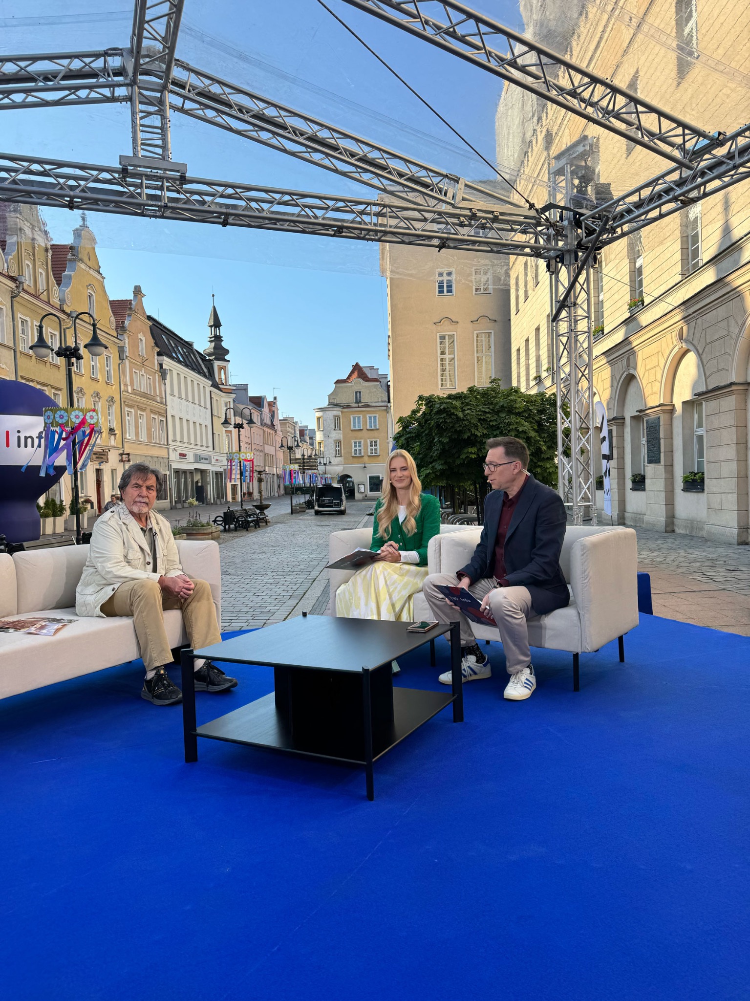 Plenerowe studio nagraniowe na tle staromiejskiej uliczki: goście siedzą na białych sofach na niebieskim dywanie pod metalową konstrukcją.