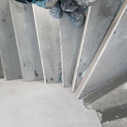 OMEGA Piotr Adamczyk - Betonowe schody w trakcie szpachlowania, widok z góry na stopy pracownika w obuwiu roboczym. Widoczne ślady zaprawy i wiadro z masą szpachlową.