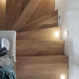 OMEGA Piotr Adamczyk - Wykończone schody z drewnopodobnych płytek, z oświetleniem punktowym w ścianie. Widoczny fragment osoby w ubraniu roboczym. Ujęcie z góry.