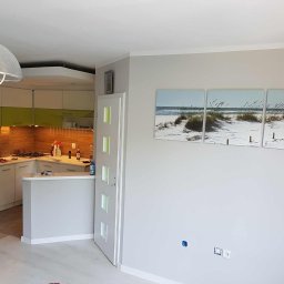 OMEGA Piotr Adamczyk - Jasne wnętrze po malowaniu: szara ściana z obrazem plaży, biała stolarka drzwiowa z zielonymi akcentami, widoczna kuchnia z zielonymi szafkami i lampą.