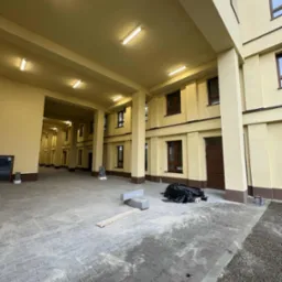 Żółty budynek z kolumnami i szarą kostką brukową. Widoczne okna z brązowymi ramami i elementy wykończeniowe. Na ziemi leżą materiały budowlane.