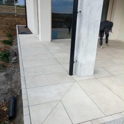 OMEGA Piotr Adamczyk - Nowoczesny taras z jasnych, kwadratowych płyt. Widoczny fragment elewacji z oknem i filarem. Osoba w tle wykonuje prace wykończeniowe przy filarze.