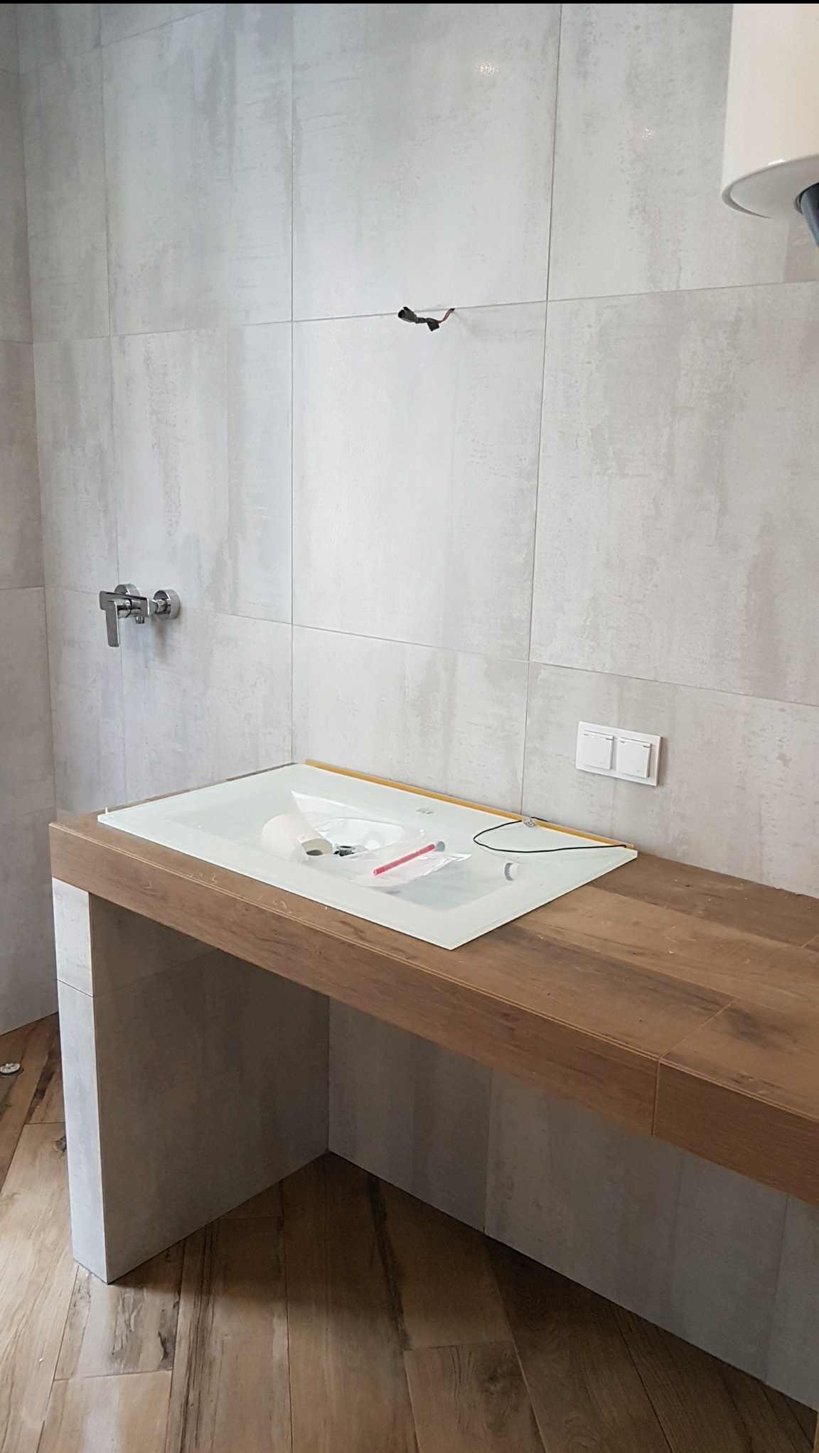 Montaż umywalki nablatowej w łazience z szarymi płytkami i drewnianym blatem. Widoczne akcesoria montażowe i instalacja elektryczna.