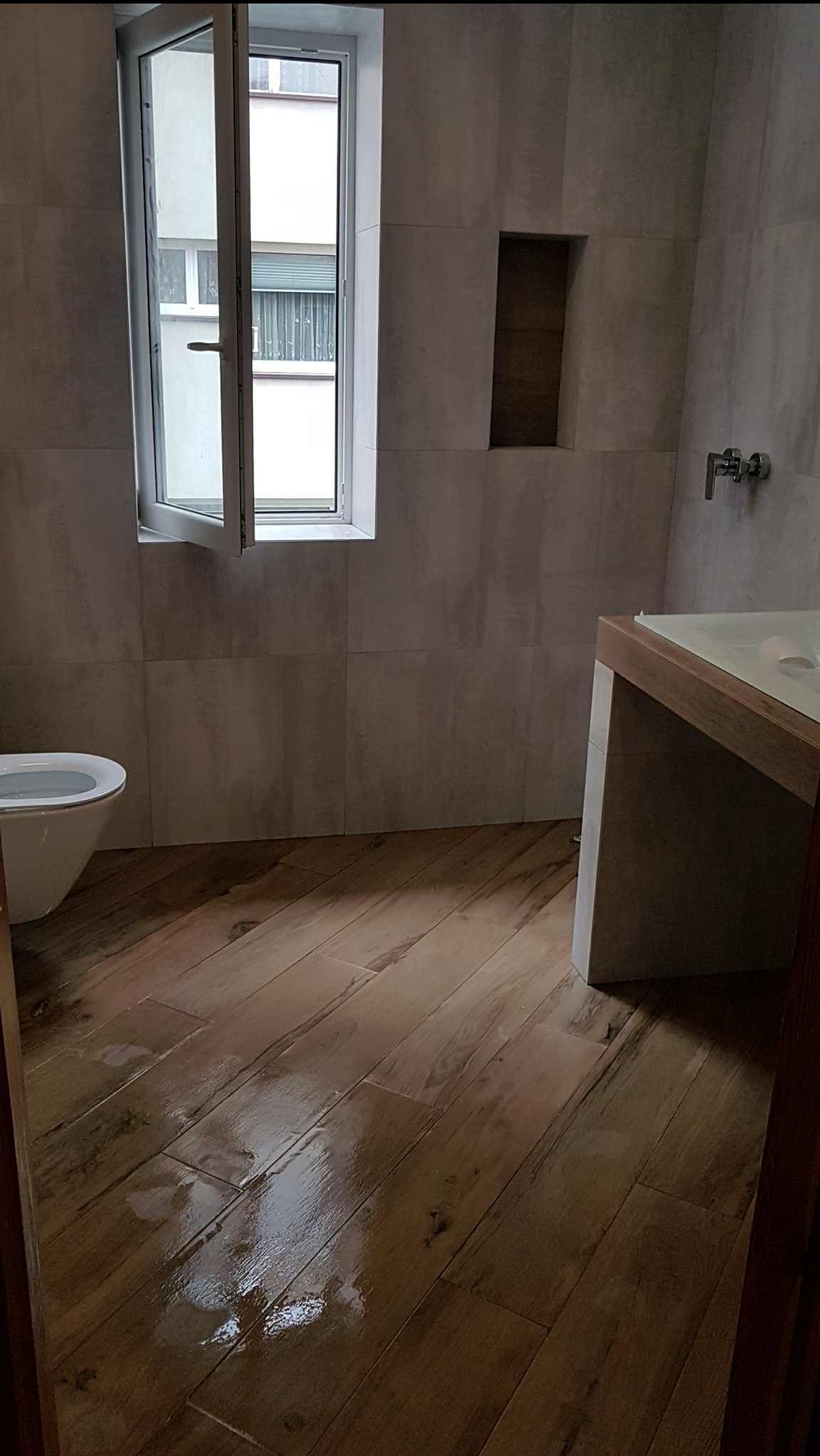 Wykończona łazienka z drewnopodobną podłogą, szarą okładziną ścian, wnęką i otwartym oknem. Widoczne elementy armatury i toaleta. Ujęcie z niskiego kąta.