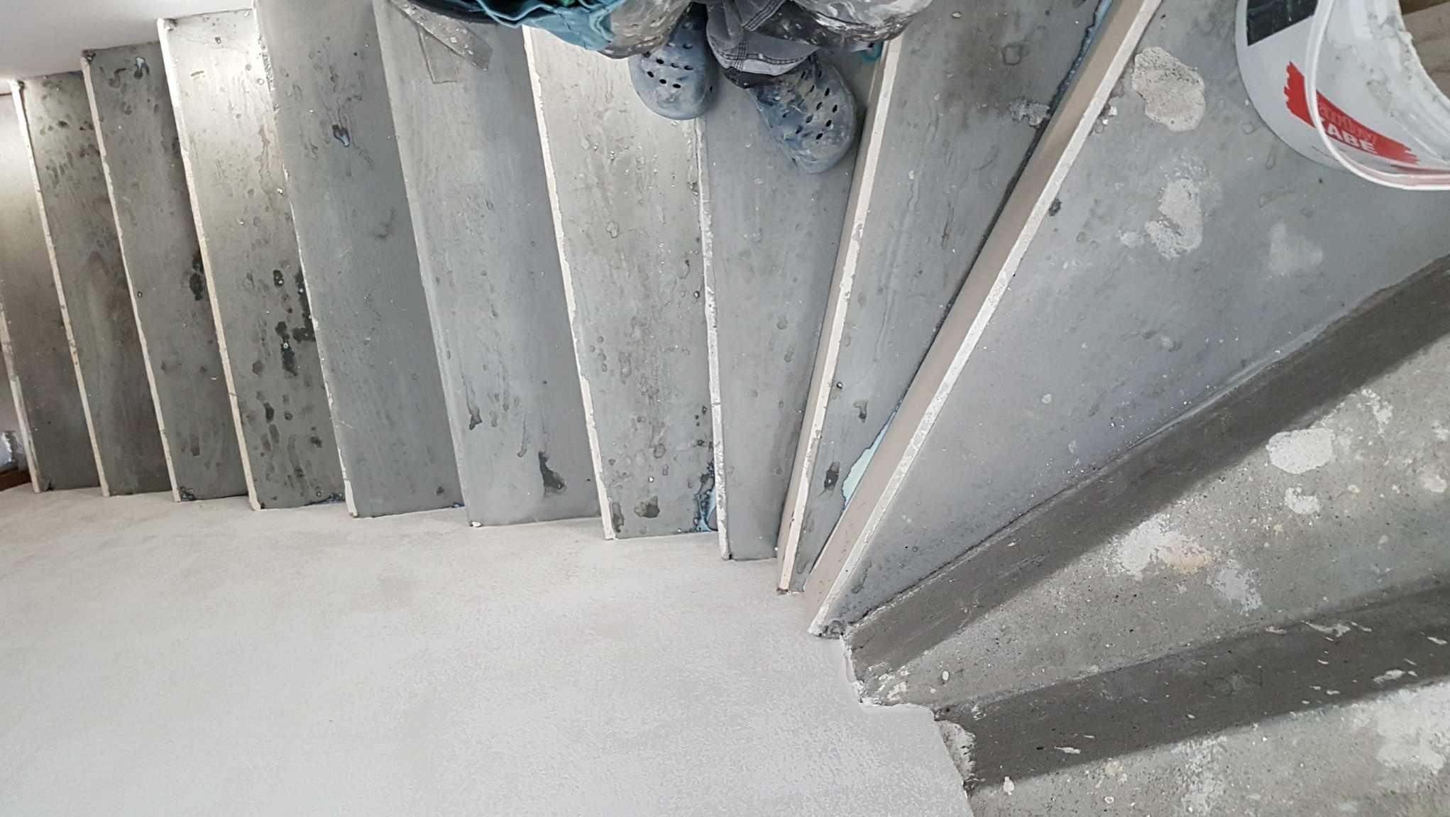 Betonowe schody w trakcie szpachlowania, widok z góry na stopy pracownika w obuwiu roboczym. Widoczne ślady zaprawy i wiadro z masą szpachlową.