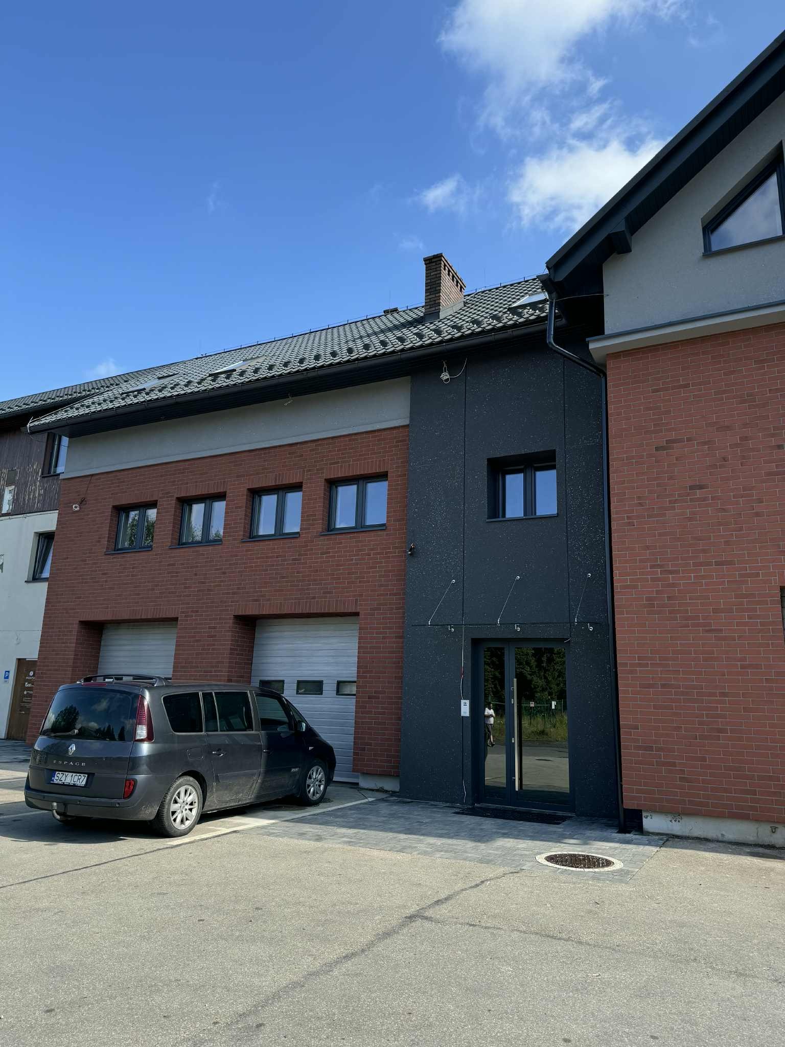 Budynek z ciemną elewacją i ceglaną fasadą, z szarym samochodem zaparkowanym przed garażem. Widoczne okna i drzwi wejściowe.