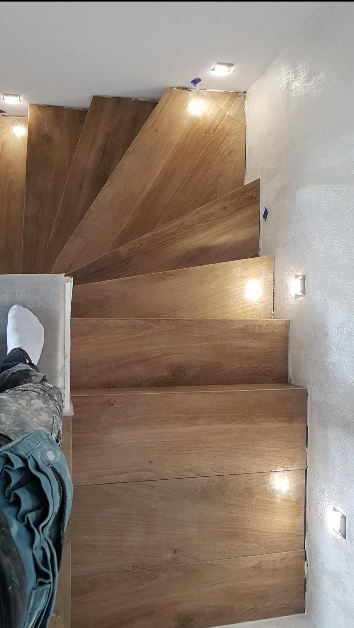 Wykończone schody z drewnopodobnych płytek, z oświetleniem punktowym w ścianie. Widoczny fragment osoby w ubraniu roboczym. Ujęcie z góry.