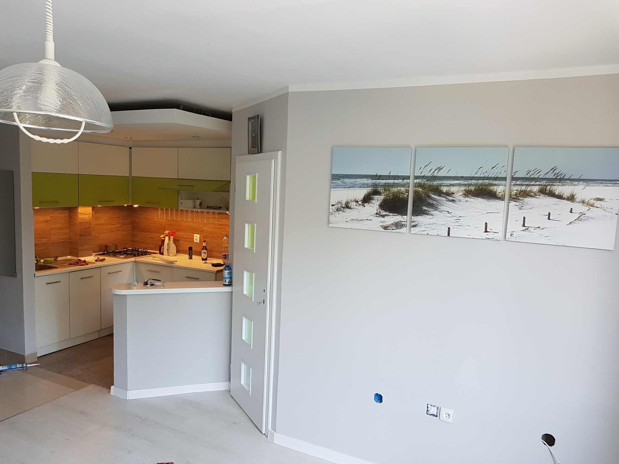 Jasne wnętrze po malowaniu: szara ściana z obrazem plaży, biała stolarka drzwiowa z zielonymi akcentami, widoczna kuchnia z zielonymi szafkami i lampą.