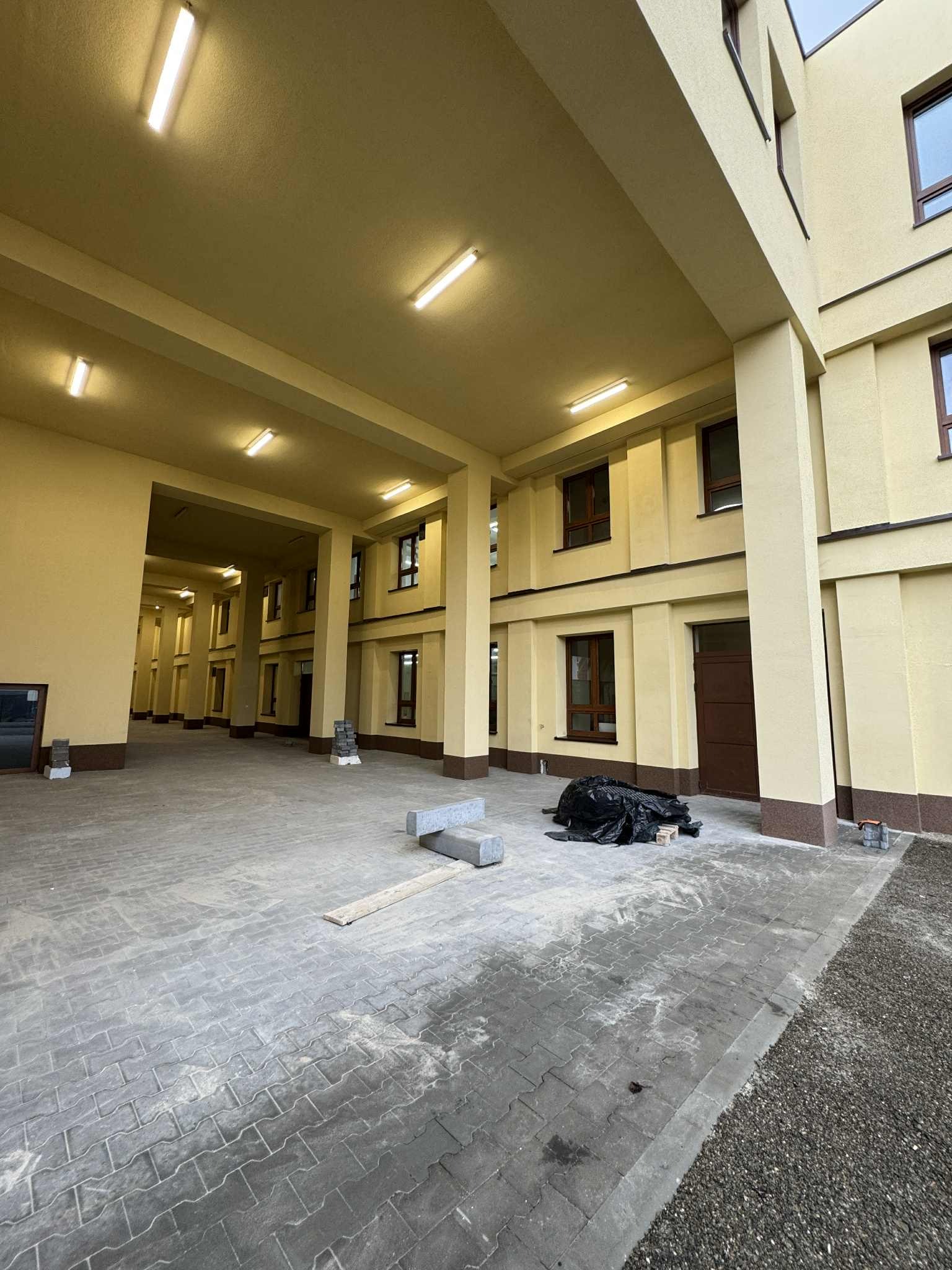 Żółty budynek z kolumnami i szarą kostką brukową. Widoczne okna z brązowymi ramami i elementy wykończeniowe. Na ziemi leżą materiały budowlane.