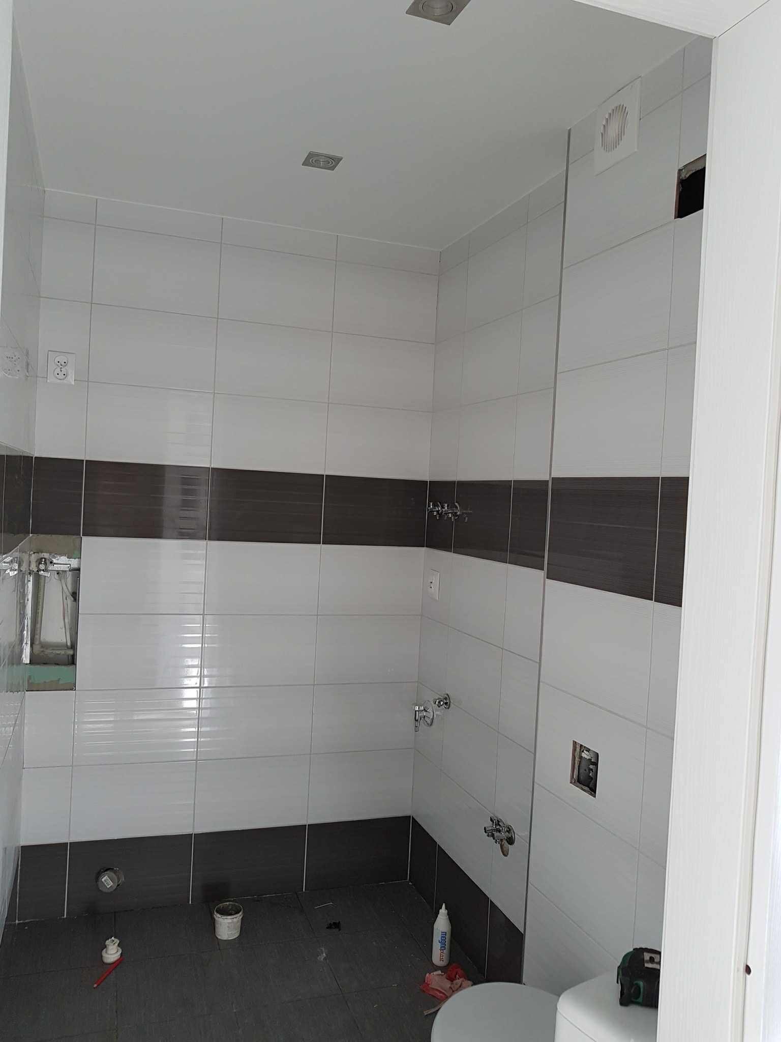 Wnętrze łazienki w trakcie prac wykończeniowych: ułożone płytki w odcieniach szarości i bieli, widoczne narzędzia glazurnicze, brakujące elementy wyposażenia.