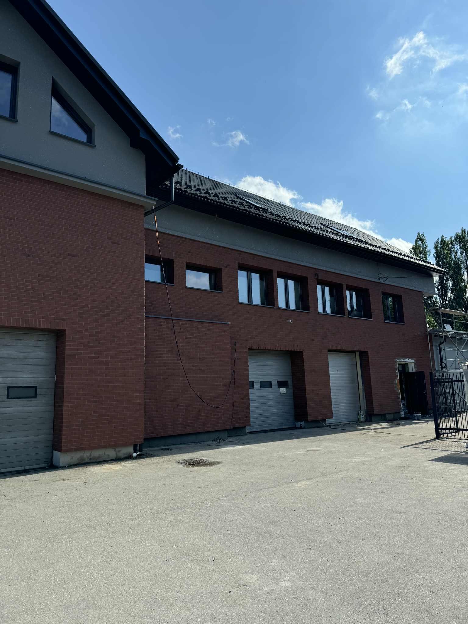 Dwukondygnacyjny budynek z cegły klinkierowej, garaże z bramami segmentowymi, okna w ciemnych ramach, szara elewacja górnej części i błękitne niebo.