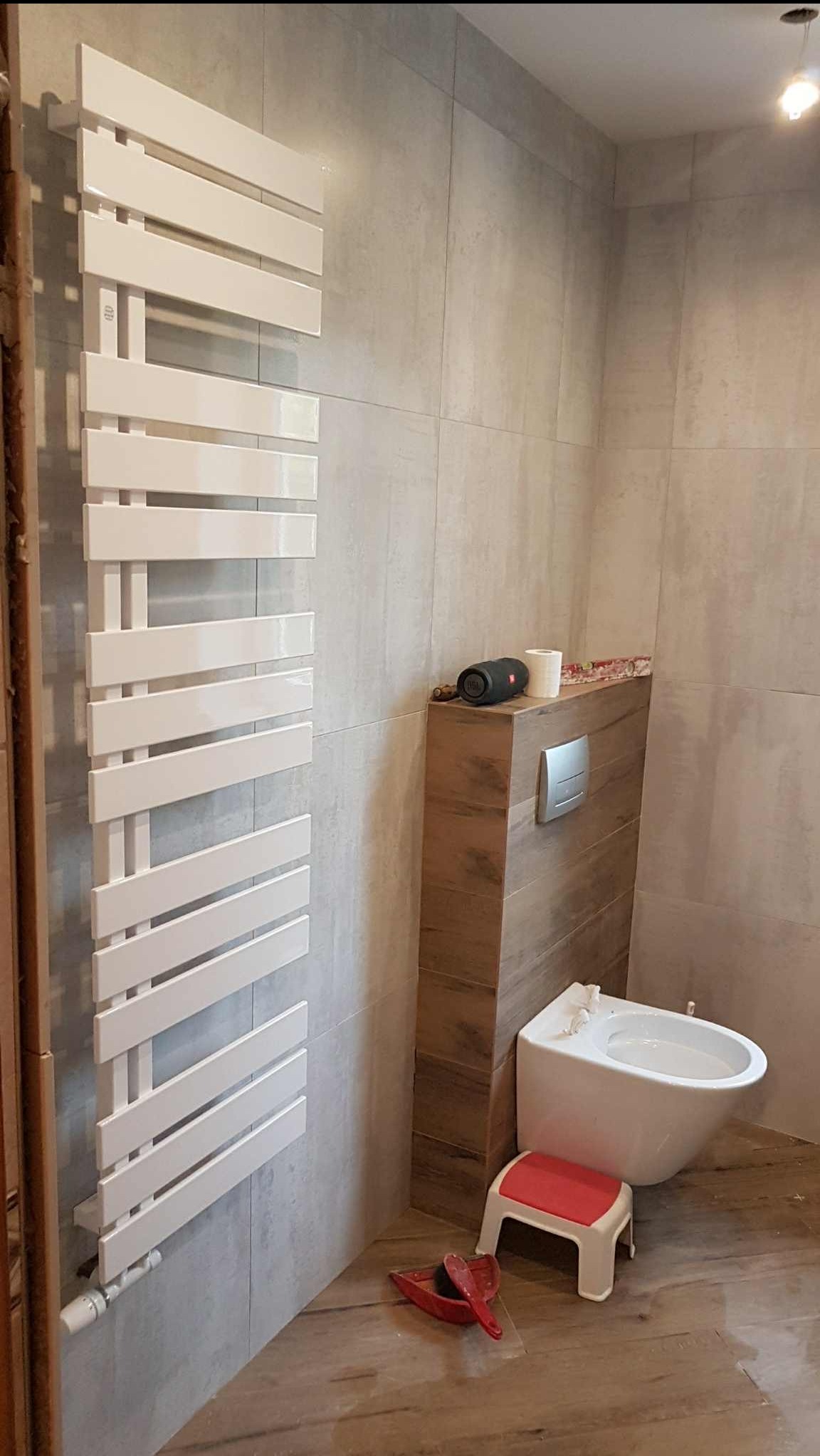 Nowoczesna łazienka z grzejnikiem drabinkowym, bidetem i szarymi płytkami. Widoczne akcesoria łazienkowe i fragment podłogi imitującej drewno.