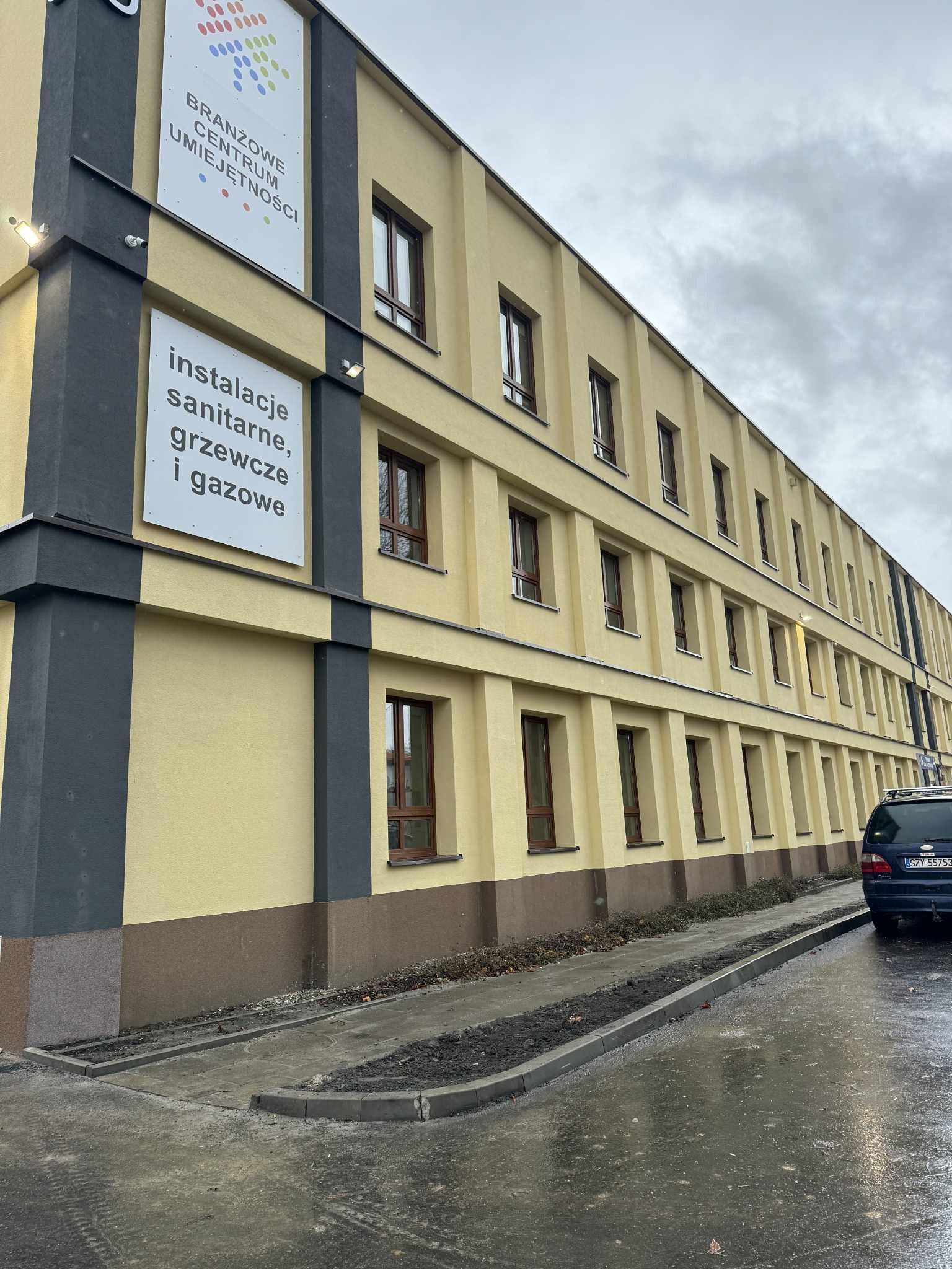 Żółty budynek z brązowymi oknami i szarymi elementami elewacji. Tablica informacyjna z napisem 'instalacje sanitarne, grzewcze i gazowe'. Pochmurne niebo.
