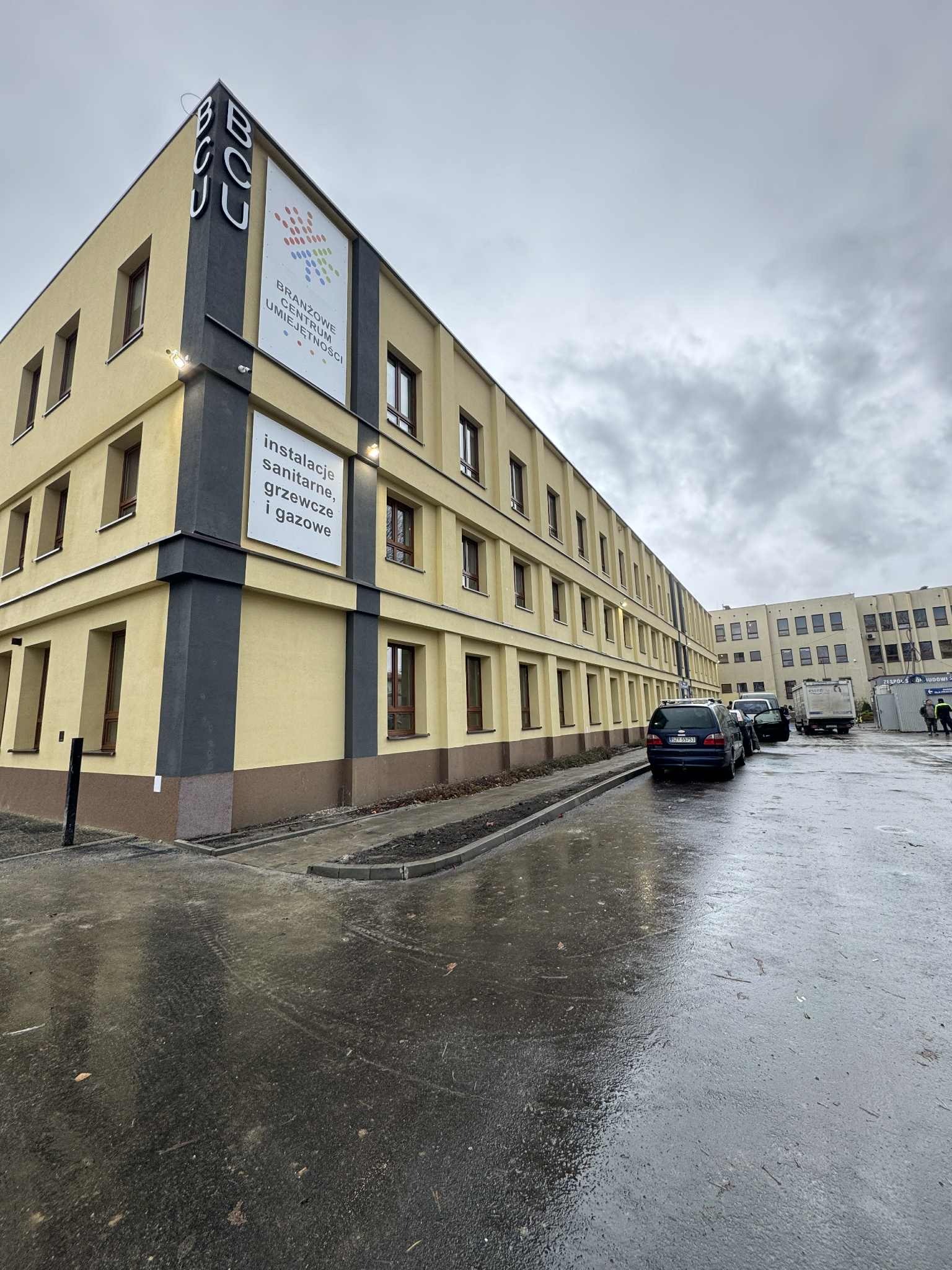 Żółty budynek BCU z szarym akcentem, tablicami informacyjnymi o instalacjach sanitarnych, grzewczych i gazowych. Mokry parking przed budynkiem, pochmurne niebo.