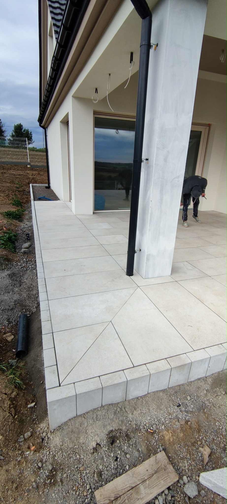 Nowoczesny taras z jasnych, kwadratowych płyt. Widoczny fragment elewacji z oknem i filarem. Osoba w tle wykonuje prace wykończeniowe przy filarze.
