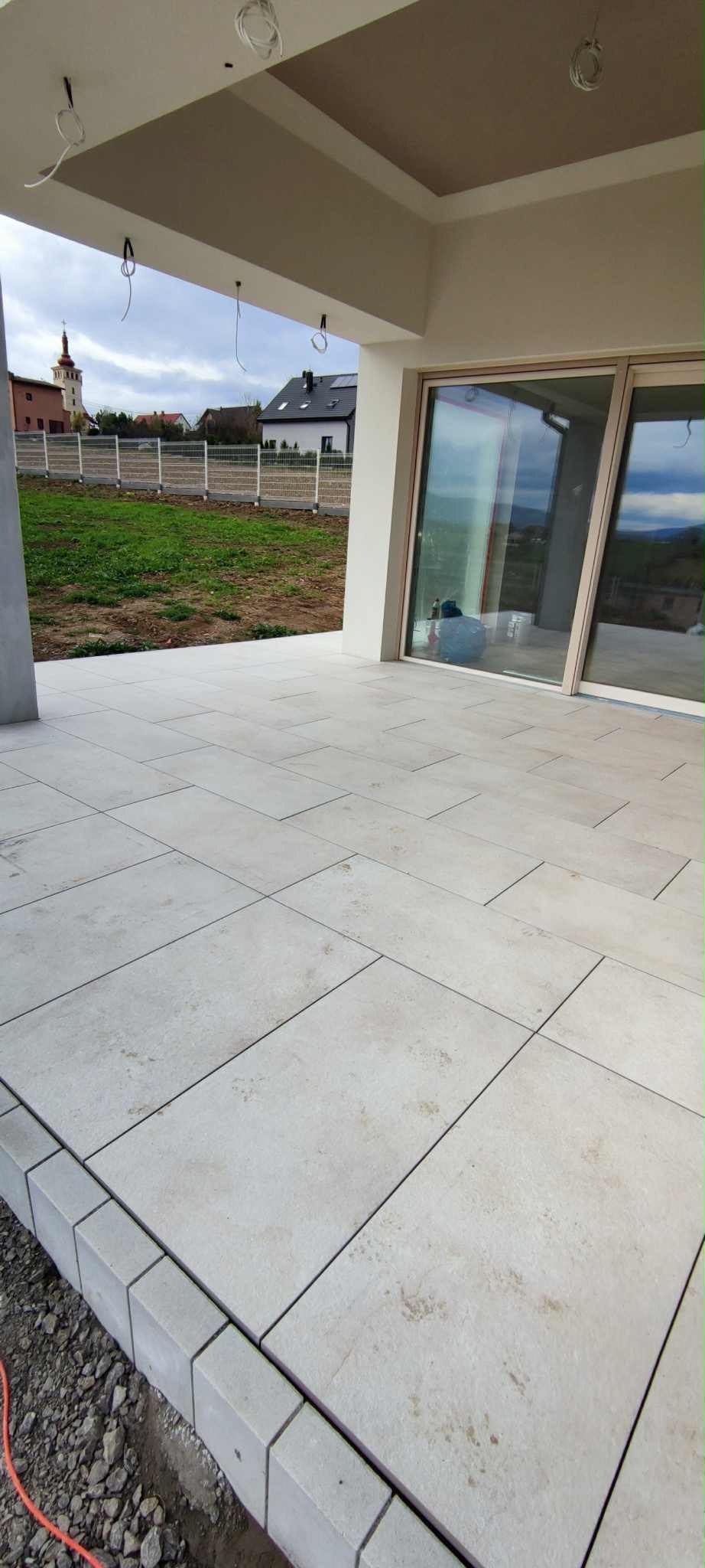 Wyłożony jasnymi płytkami taras z widokiem na ogród i domy. Płytki ułożone równo, widoczne fugi. Częściowo widoczne kable wystające z sufitu.