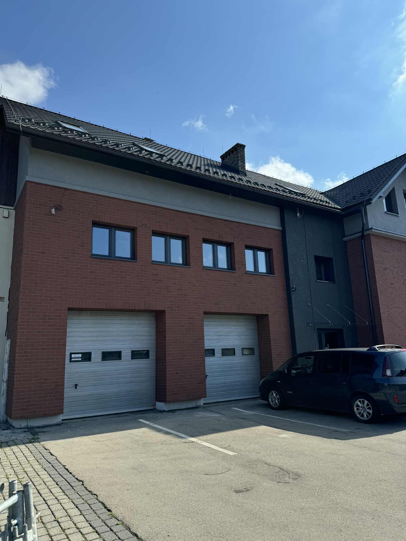 Dwukondygnacyjny budynek z dwoma garażami, wykończony cegłą i szarym tynkiem, z zaparkowanym samochodem na parkingu. Widok na elewację i dach.