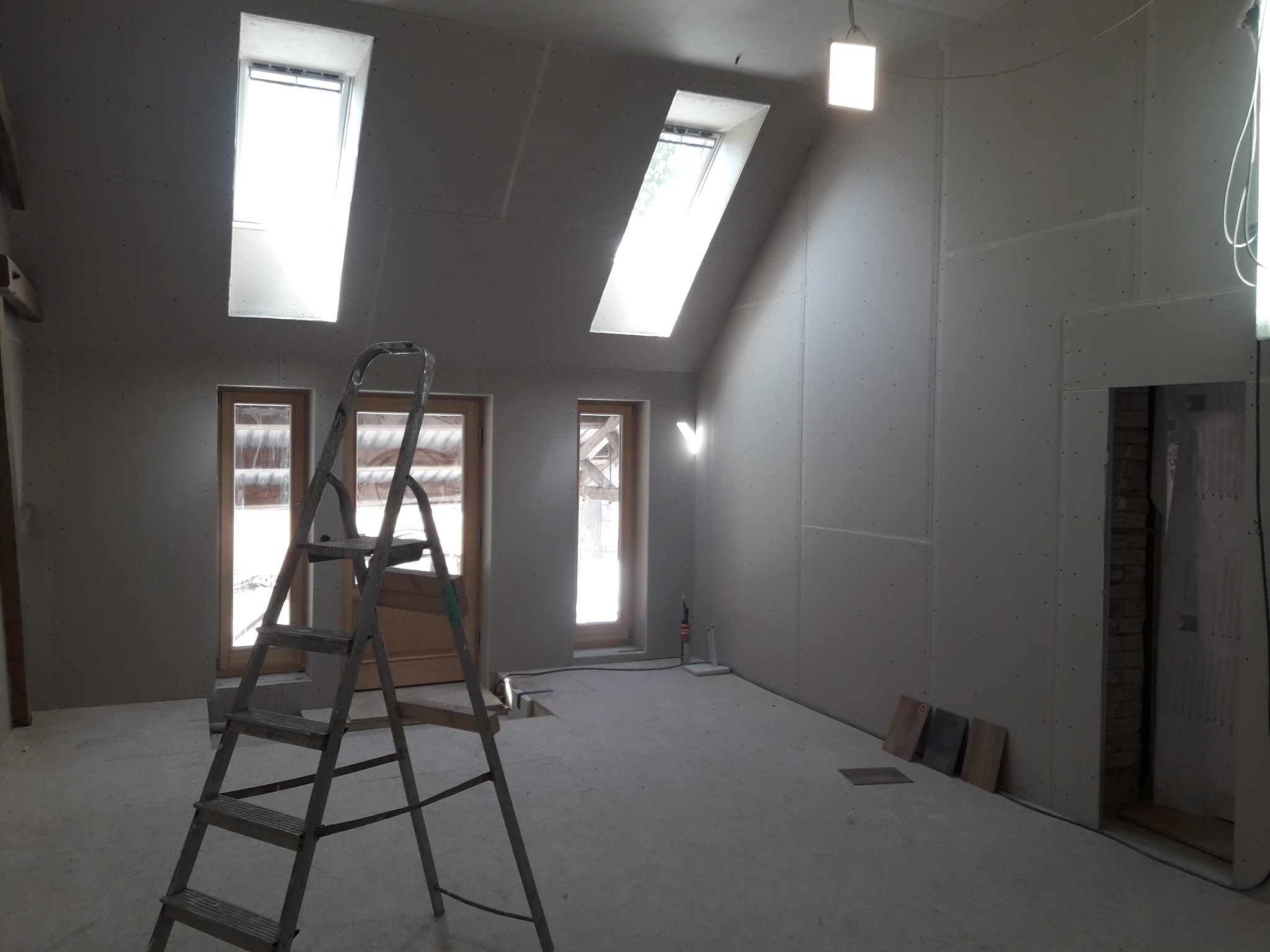 Poddasze w trakcie adaptacji: ściany wyłożone płytami karton-gips, okna dachowe, drabina, płytki pod ścianą. Widoczne przygotowania do dalszych prac wykończeniowych.