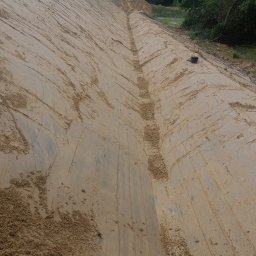 Digger-pro roboty ziemne - Szeroki wykop w ziemi z wyraźnymi śladami maszyn, prawdopodobnie przygotowany pod budowę drogi lub stawu, z lasem w tle i pochmurnym niebem.