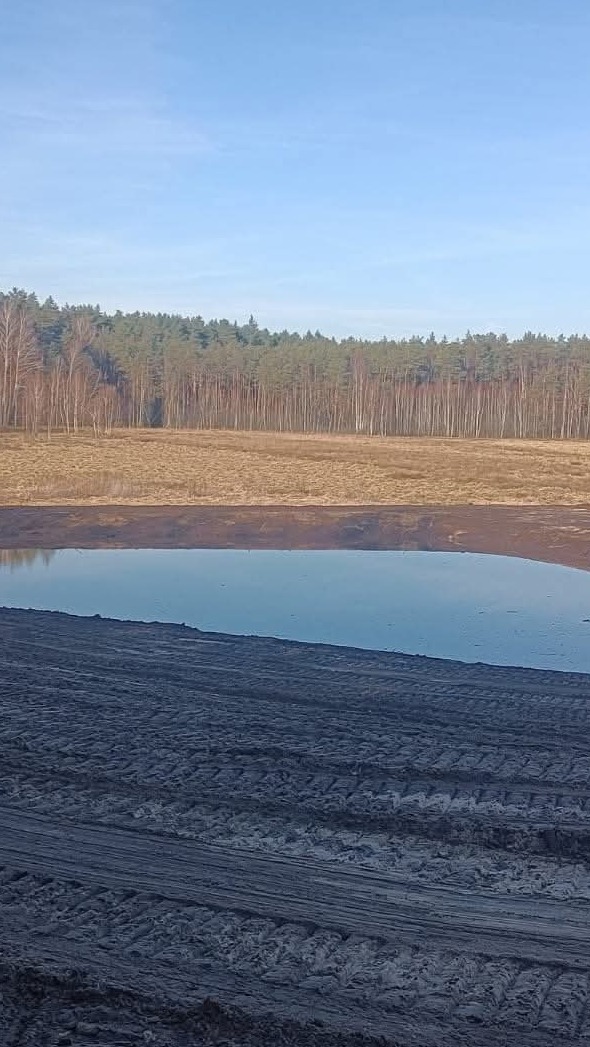 Widok na częściowo zalany teren z widocznymi śladami opon, w tle las i błękitne niebo. Teren przygotowany pod inwestycję.