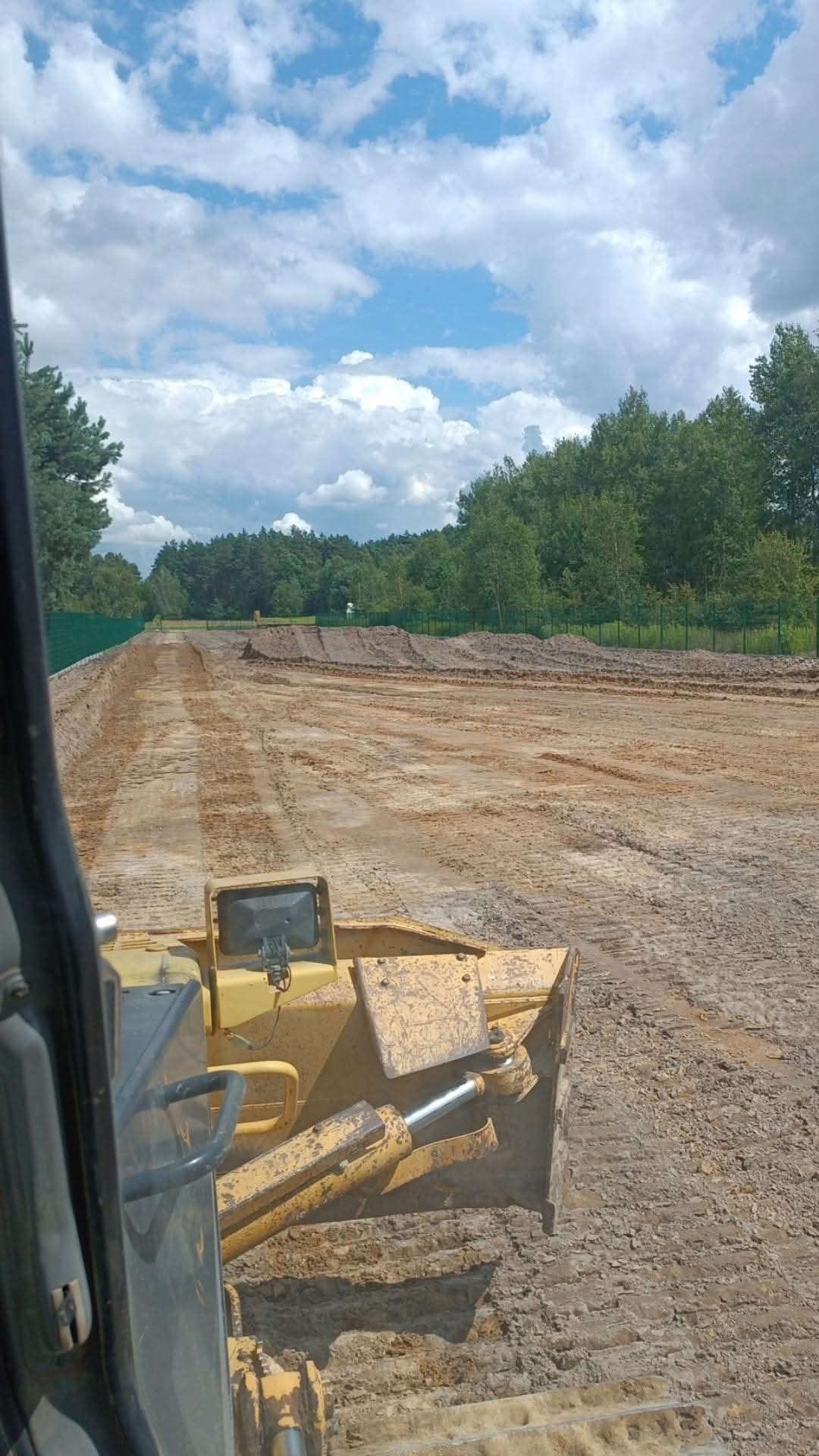 Żółty spychacz w trakcie niwelacji terenu, widok z kabiny operatora. W tle widoczne drzewa i błękitne niebo z obłokami.