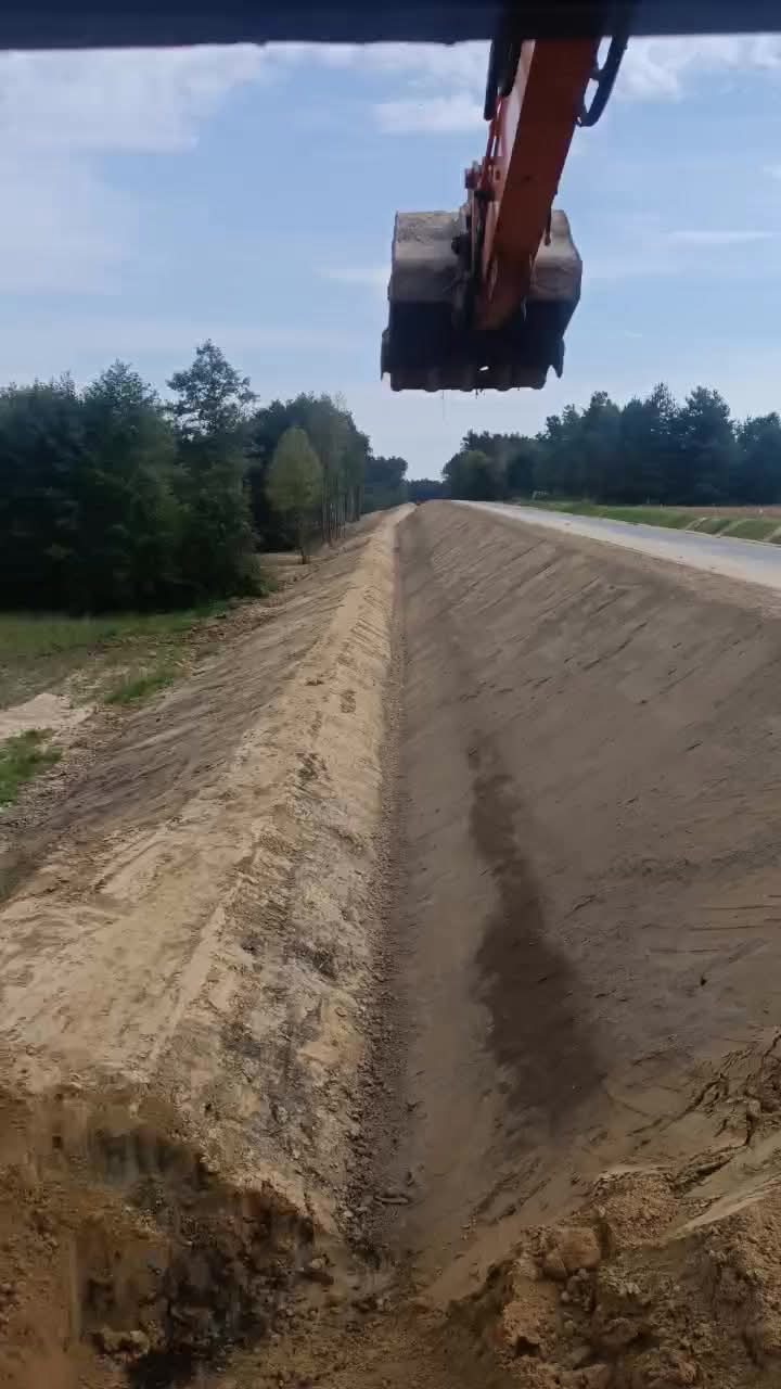 Pomarańczowa koparka nad wykopanym rowem w ziemi, widok z góry, w tle drzewa i niebo. Prace ziemne w toku, równanie terenu.