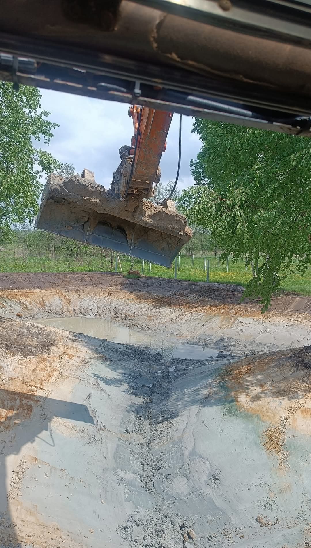 Pomarańczowa koparka z łyżką nad wykopanym stawem, widok z kabiny. W tle zielona łąka i drzewa, błękitne niebo. Woda w stawie odbija niebo.