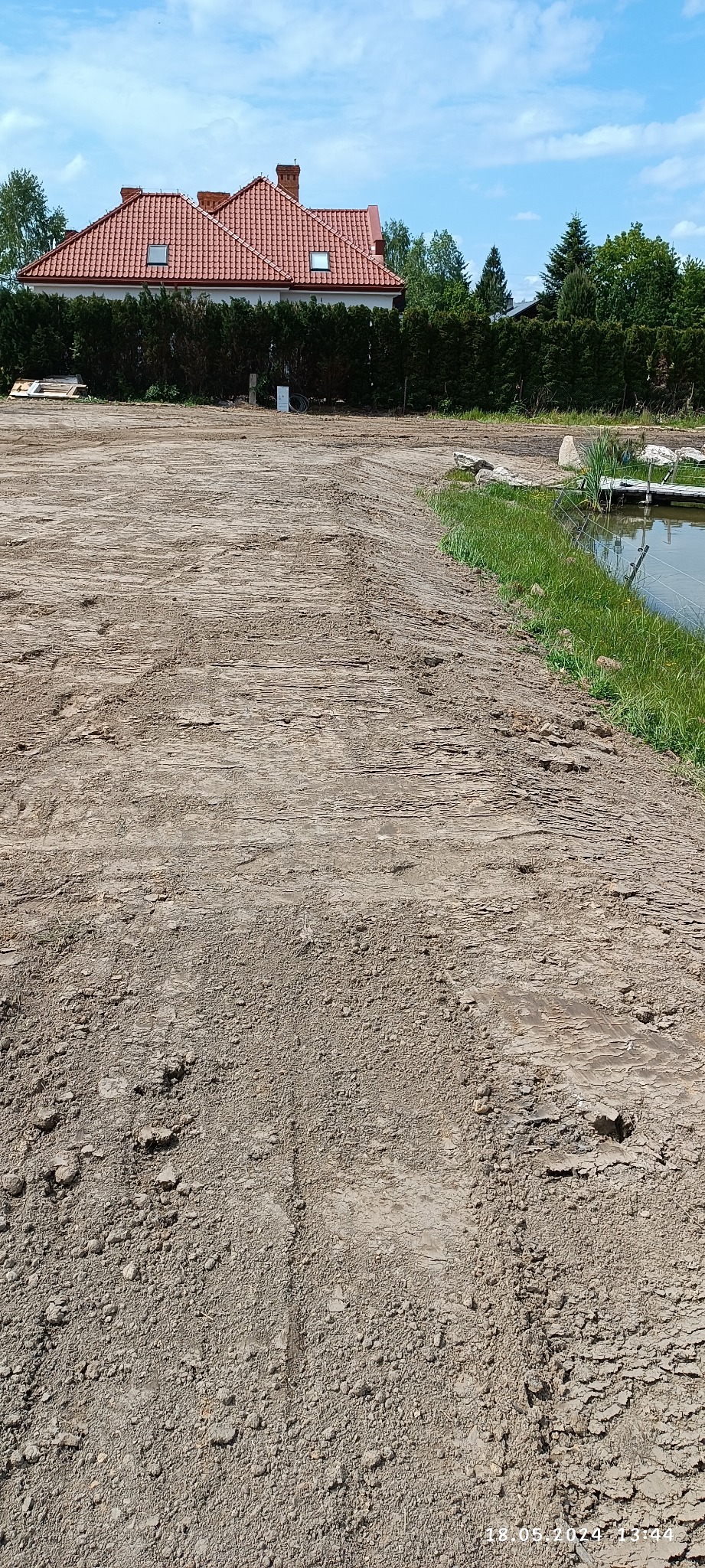 Ukształtowany teren z widocznymi śladami maszyn, przygotowany pod budowę domu z czerwonym dachem. Z boku staw z pomostem i zielenią.