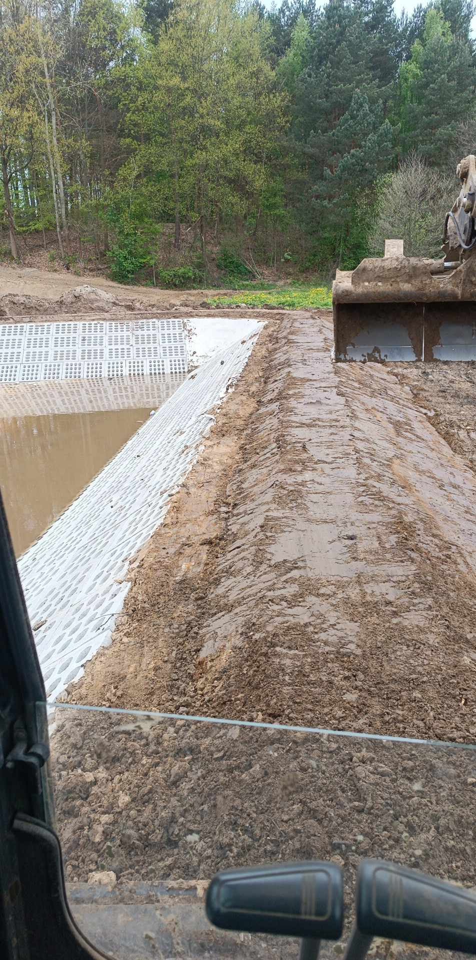 Wykopany staw z umocnionymi brzegami z betonowych elementów. W tle drzewa, w kadrze fragment koparki. Prace ziemne w toku, widok z kabiny operatora.
