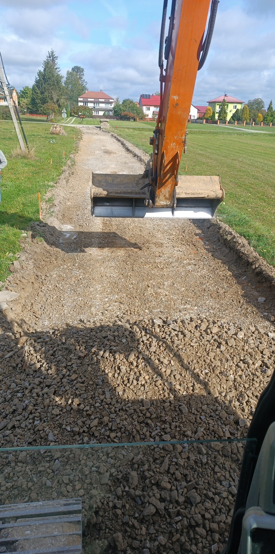 Pomarańczowa koparka rozkłada kamień na utwardzanej drodze gruntowej. Widoczne domy w tle i błękitne niebo. Na pierwszym planie kamienie.