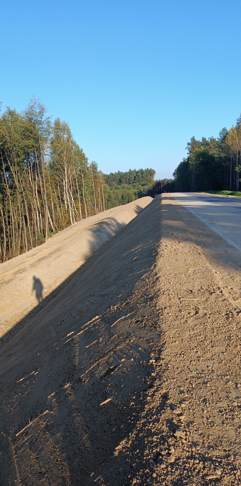 Świeżo usypany nasyp ziemny wzdłuż drogi, widoczne ślady opon i cień osoby, drzewa w tle pod błękitnym niebem.