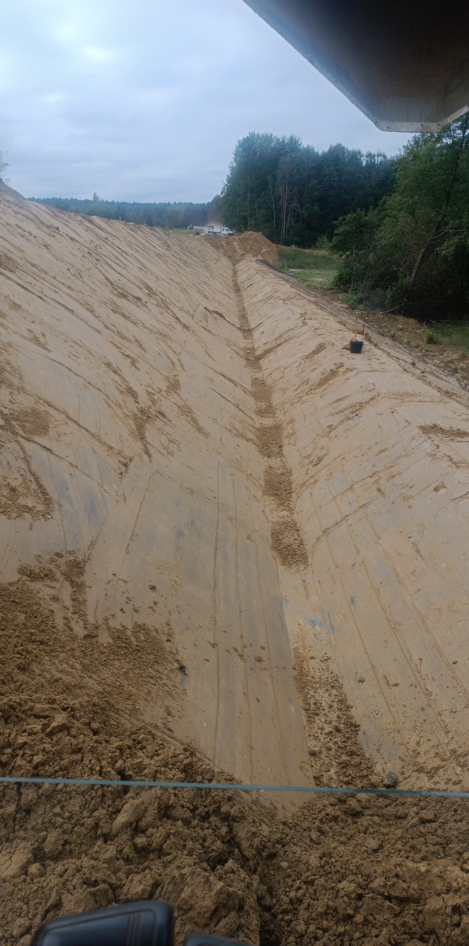Szeroki wykop w ziemi z wyraźnymi śladami maszyn, prawdopodobnie przygotowany pod budowę drogi lub stawu, z lasem w tle i pochmurnym niebem.
