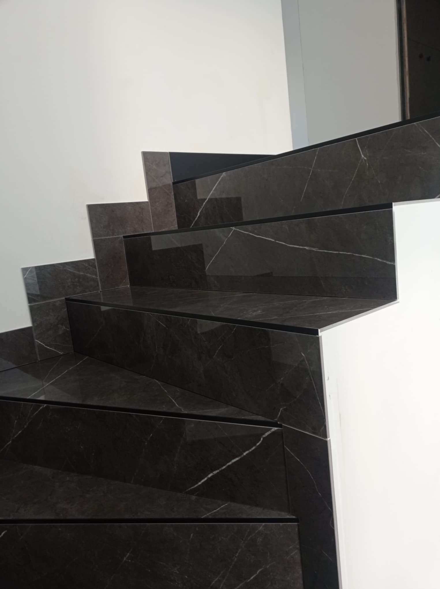 Wykończone schody z ciemnego marmuru z białymi żyłami, z czarnymi listwami wykończeniowymi, na tle białej ściany. Widok z dołu, ukazujący detale wykończenia.
