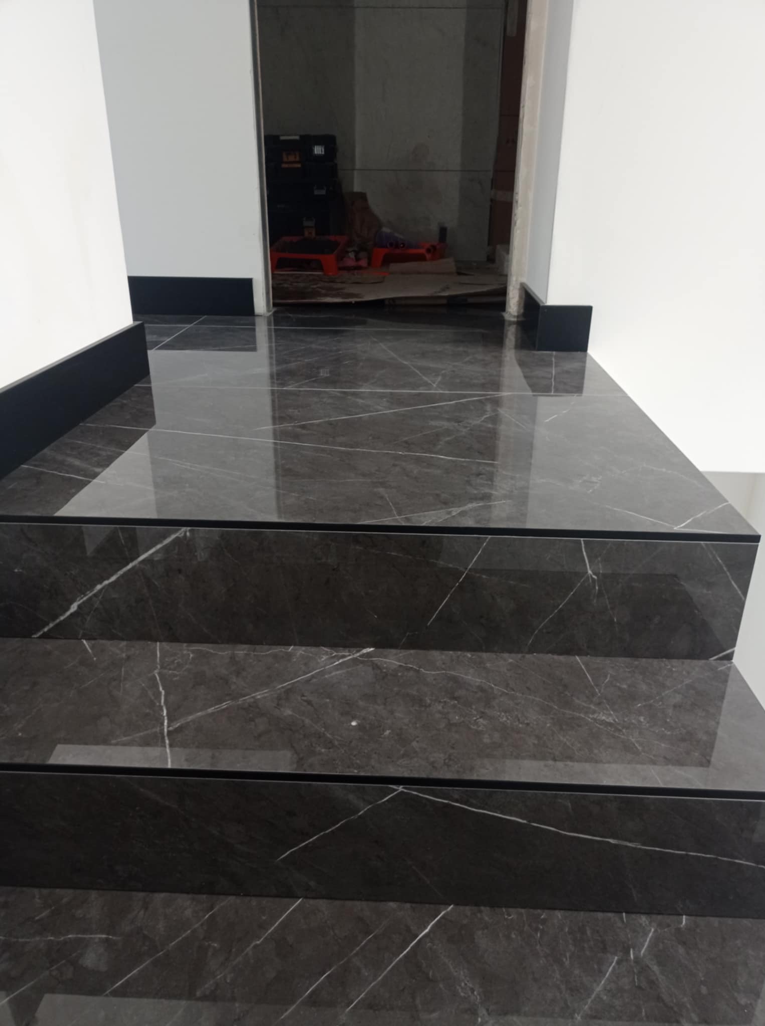 Schody wykończone ciemnym gresem imitującym marmur z białymi żyłami, widoczne z dołu, z czarnymi cokołami przy białych ścianach. W tle widoczne narzędzia.