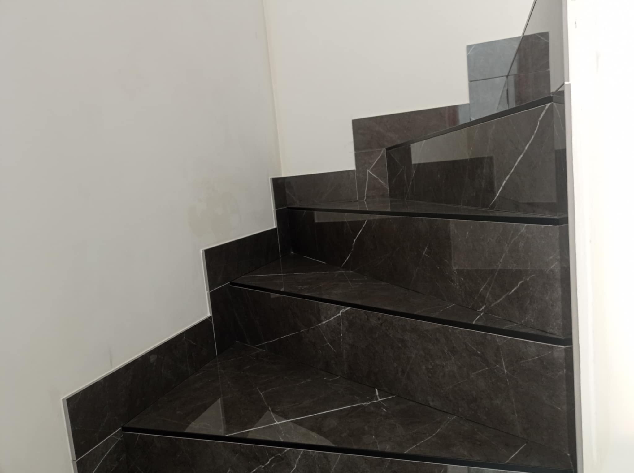 Wykończone schody z ciemnego marmuru z białymi żyłami, elegancko wkomponowane w jasne wnętrze. Widoczne detale wykończenia i precyzja ułożenia płytek.