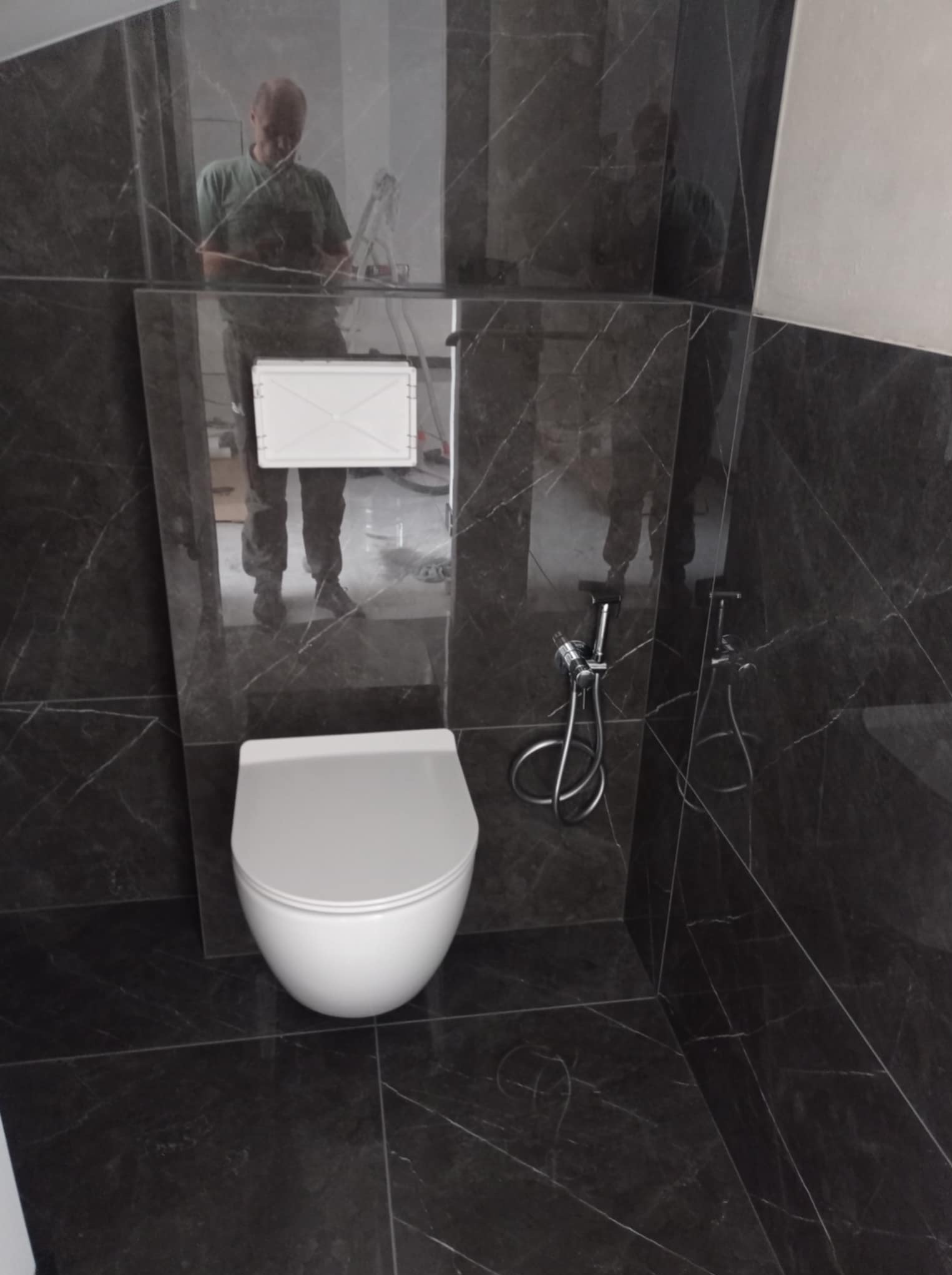 Nowoczesna toaleta w łazience wykończonej ciemnym marmurem z wyraźnym użyleniem. Widoczny biały sedes podwieszany i bidetka. W lustrzanej powierzchni odbija się osoba.